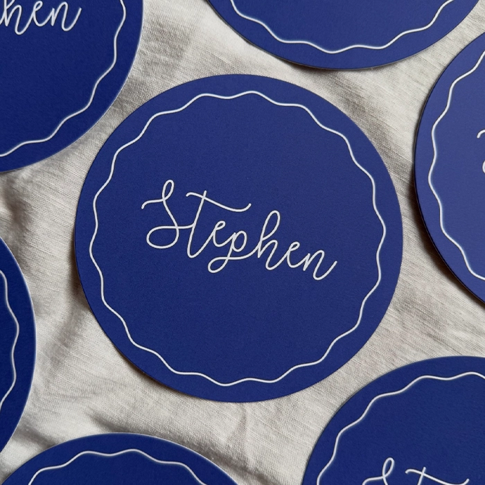 Round, navy blue, flat wedding place card with a wavy white border and the name 'Stephen' written in elegant cursive in the centre.