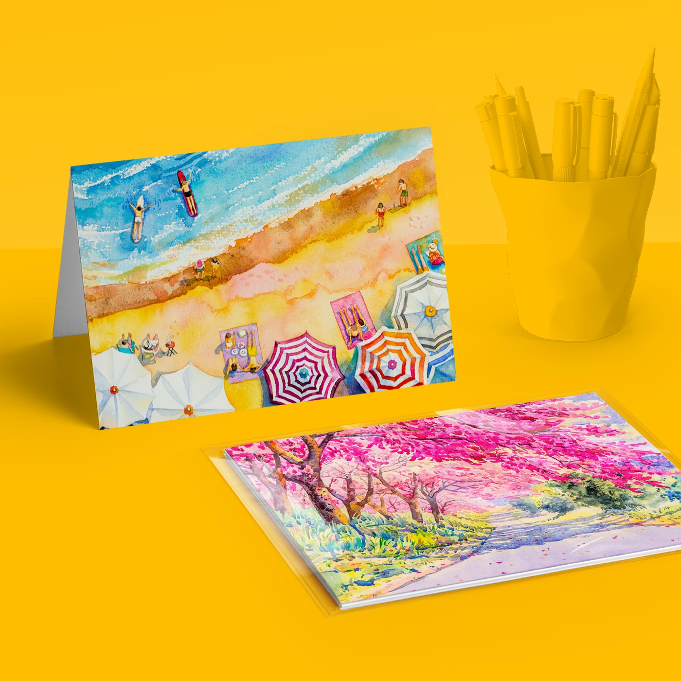 Tent fold illustrated greeting card showing a beach scene, and another greeting card inside a cellophane bag. Both on a yellow background featuring a pencil pot.