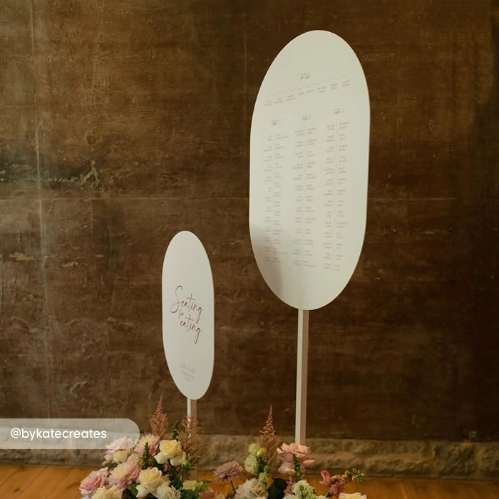 Double arch wedding seating chart signs on stands with floral arrangements at the base against a wall.
