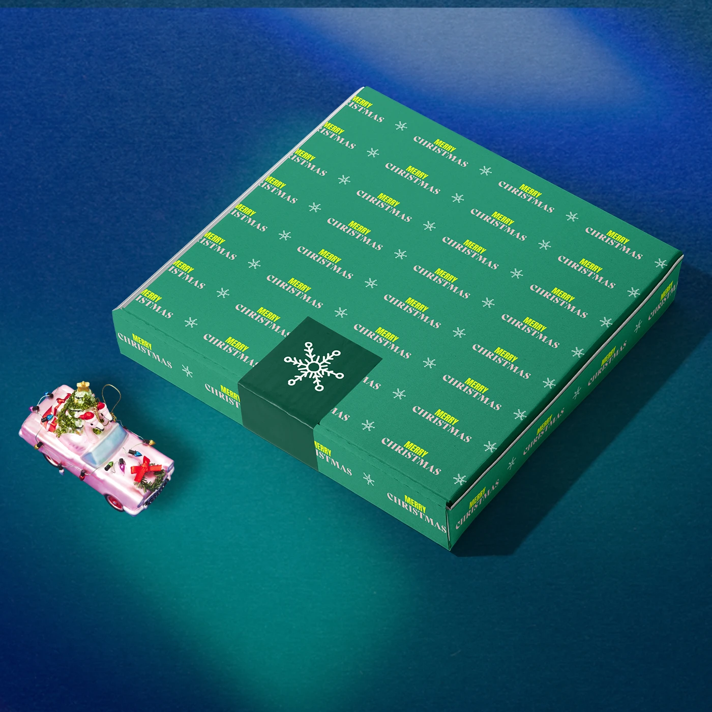 Green printed box featuring repeated 'Merry Christmas' text, on a dark blue background beside a pink christmas ornament.