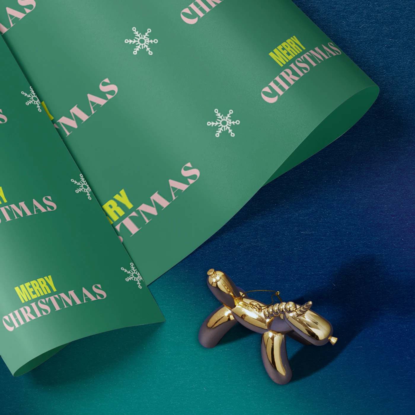 Close up image of a Wrapping Paper Sheet with a dark green design featuring repeat text reading 'Merry Christmas', on a dark blue background beside a gold Christmas ornament.