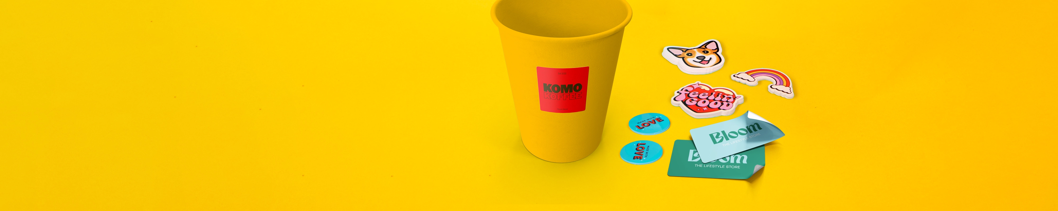 Yellow cup with a red ‘KOMO KOFFEE’ sticker surrounded by coloruful stickers including a corgi face, rainbow, ‘Feelin’ Good,’ and ‘LOVE’ designs on a bright yellow background
