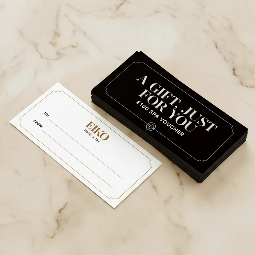 A stack of black spa gift vouchers reading “A Gift Just For You £100 Spa Voucher” next to a blank white “To/From” card on a surface.