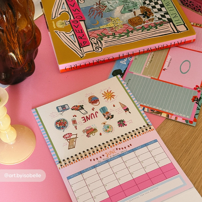 A colourful desk scene with a small open calendar showing the month of June, illustrated with cheerful drawings like flowers, an ice cream, and a sun. Next to it are patterned notepads, a decorative book with a bright cover, and a glass candle holder.