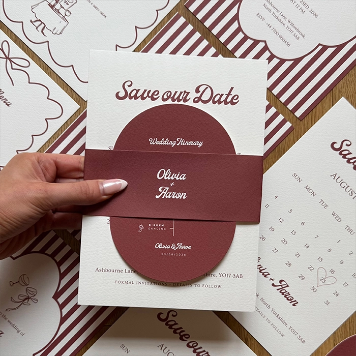Hand holding a burgundy-and-cream “Save our Date” wedding card with a wrap band reading “Olivia + Aaron,” set over matching stationery in the background.