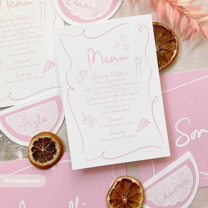 Blush-pink wedding Menu Card with handwritten-style text, flat-laid among matching place cards and dried citrus slices on a light background.