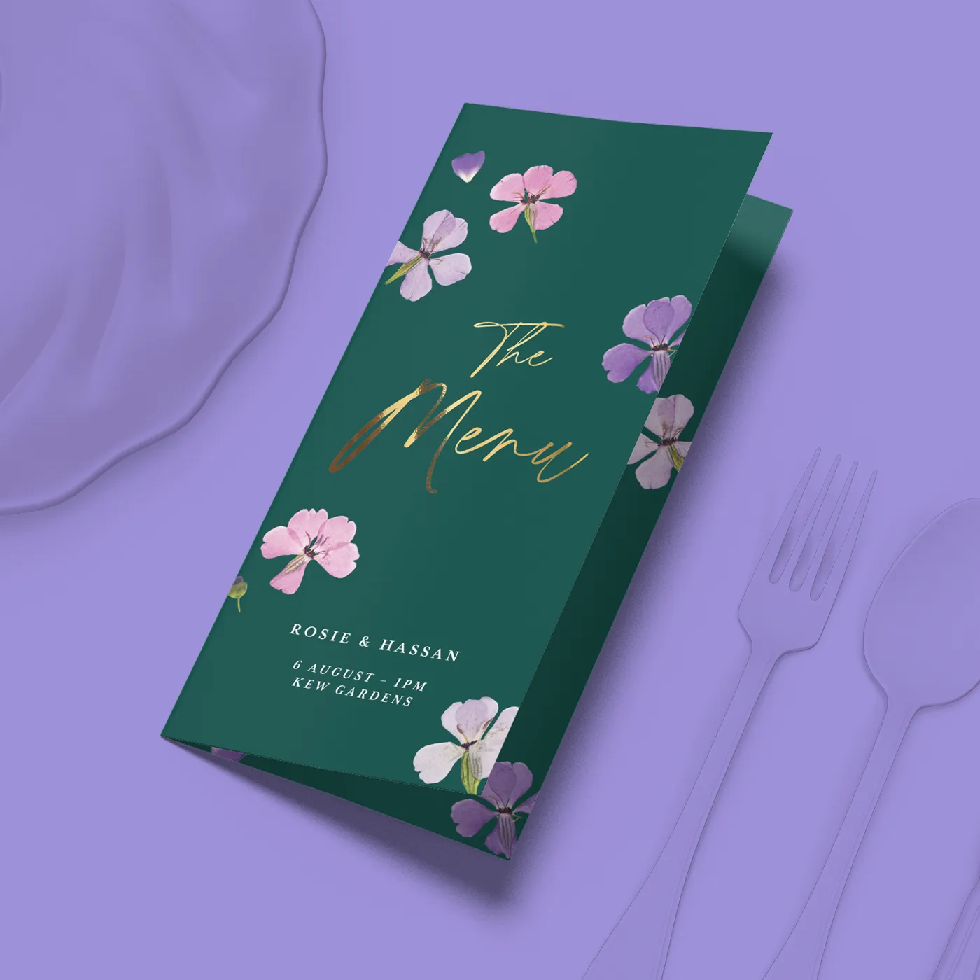 Folded green menu card with pink and purple florals, displayed on a lavender surface with plate and cutlery.