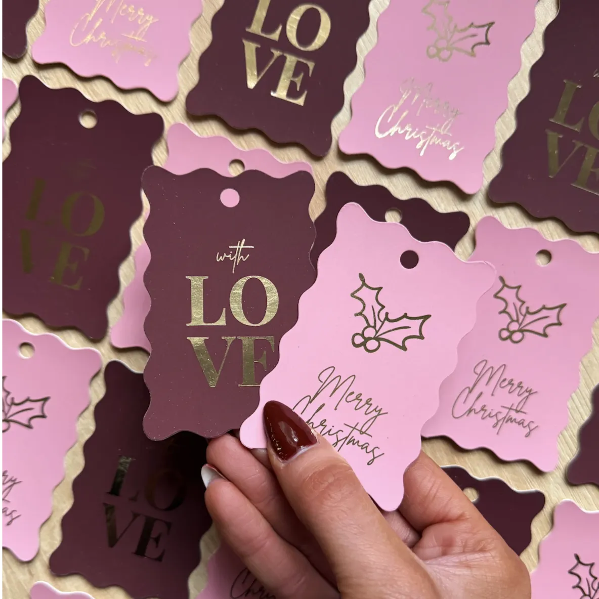 Hand holding a scalloped-edge pink Gift Tag with “Merry Christmas” in cursive and a holly illustration. Surrounding tags include maroon versions with gold foil “LOVE” text and matching scalloped edges, all designed for festive holiday gifting