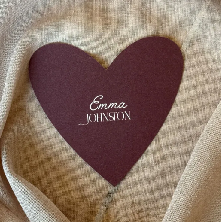 Heart-shaped maroon Wedding Place Card featuring “Emma Johnson” in white mixed-style lettering, styled on soft beige fabric