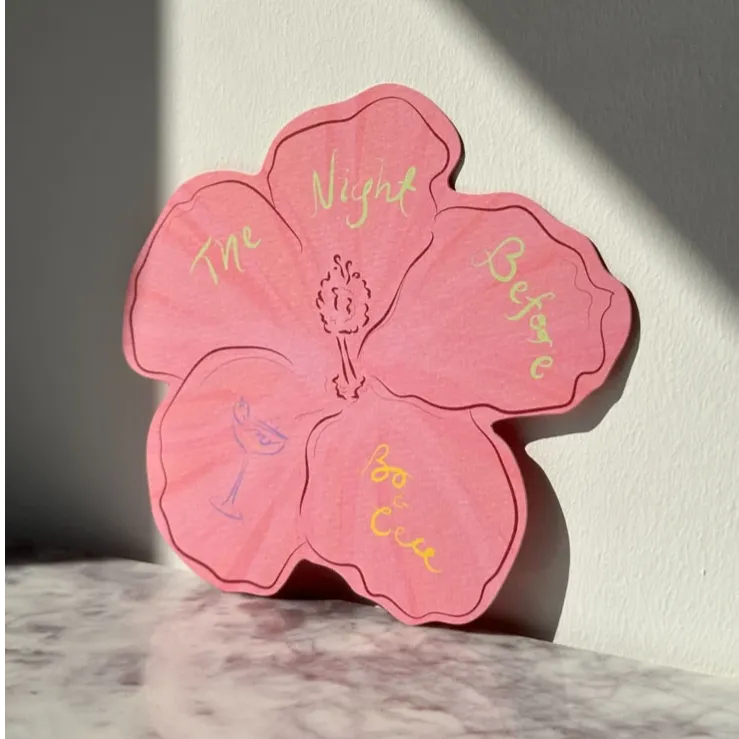 Pink flower-shaped wedding card with pastel text reading “The Night Before” placed on marble.