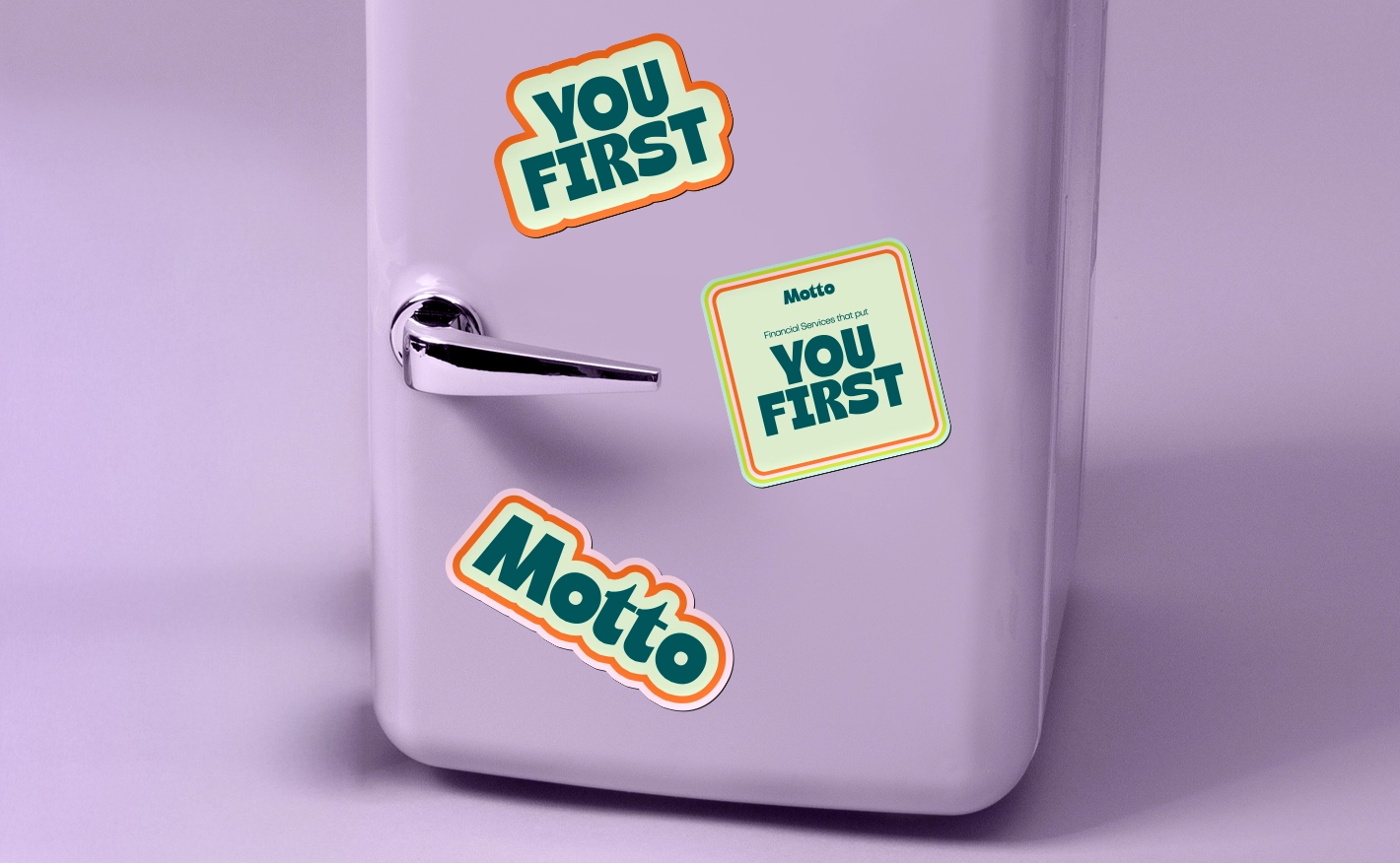 Three custom shaped magnets on a light purple refrigerator door: “YOU FIRST” in bold green and orange text, a square shape reading “Motto YOU FIRST” with a blue border, and a smaller “Motto” magnet in orange and green.