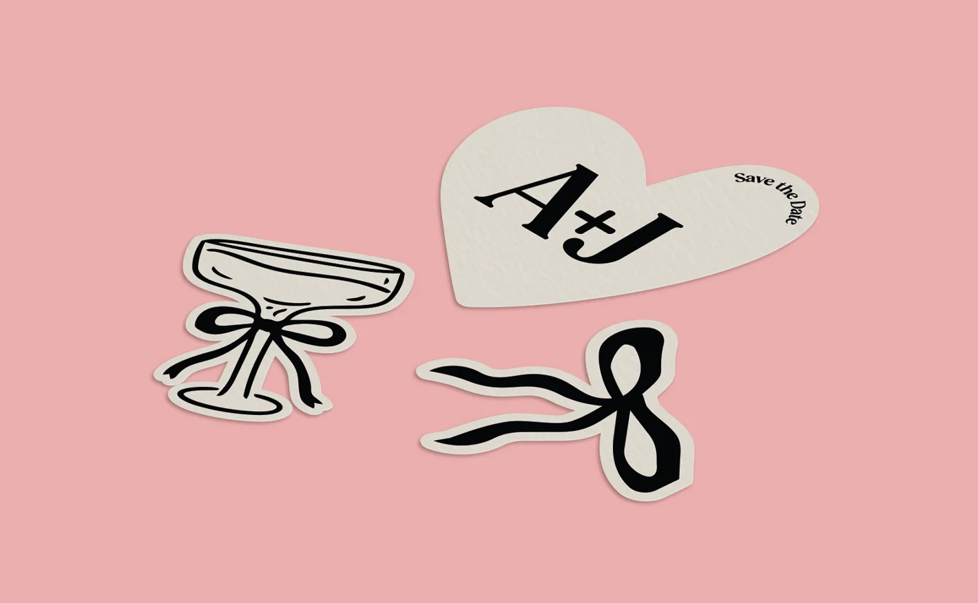 Three decorative custom shaped prints on a pink background: a stylised champagne glass with a black ribbon, a standalone black bow, and a white heart-shaped piece with bold “A + J” text and a small “save the date” note.