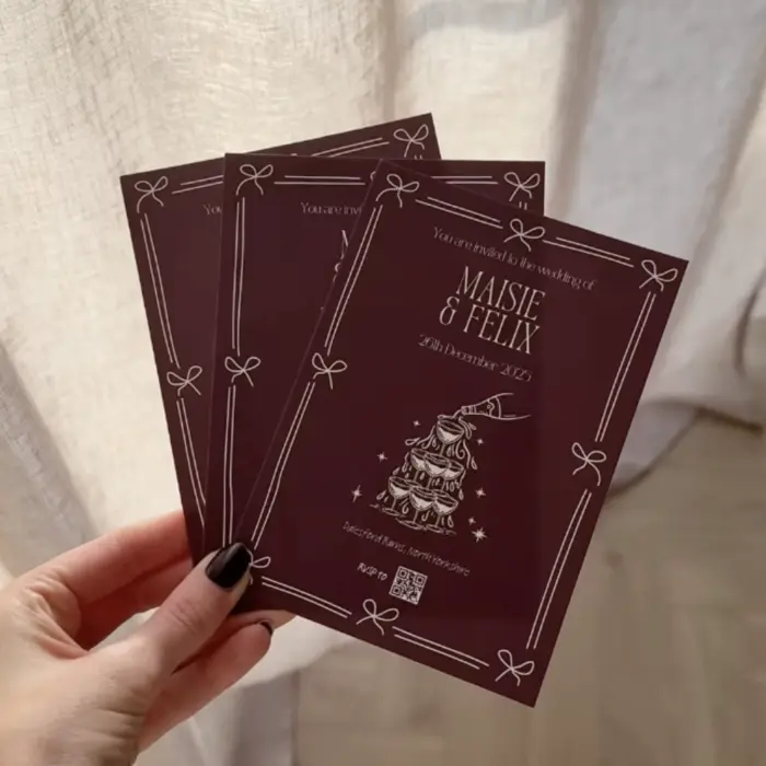A hand holds three identical burgundy wedding invitations featuring a white illustrated tiered cake topped with a wizard hat. The cards read “You’re invited to the wedding of Maine & Felix, 23 December 2023, Location: Hogwarts Great Hall” with a QR code at the bottom.