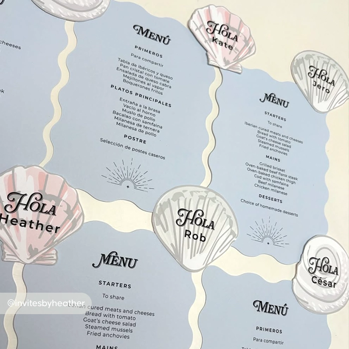 A flat lay of blue wavy Wedding Menus paired with seashell‑shaped name cards. The Menus list starters, mains, and dessert.