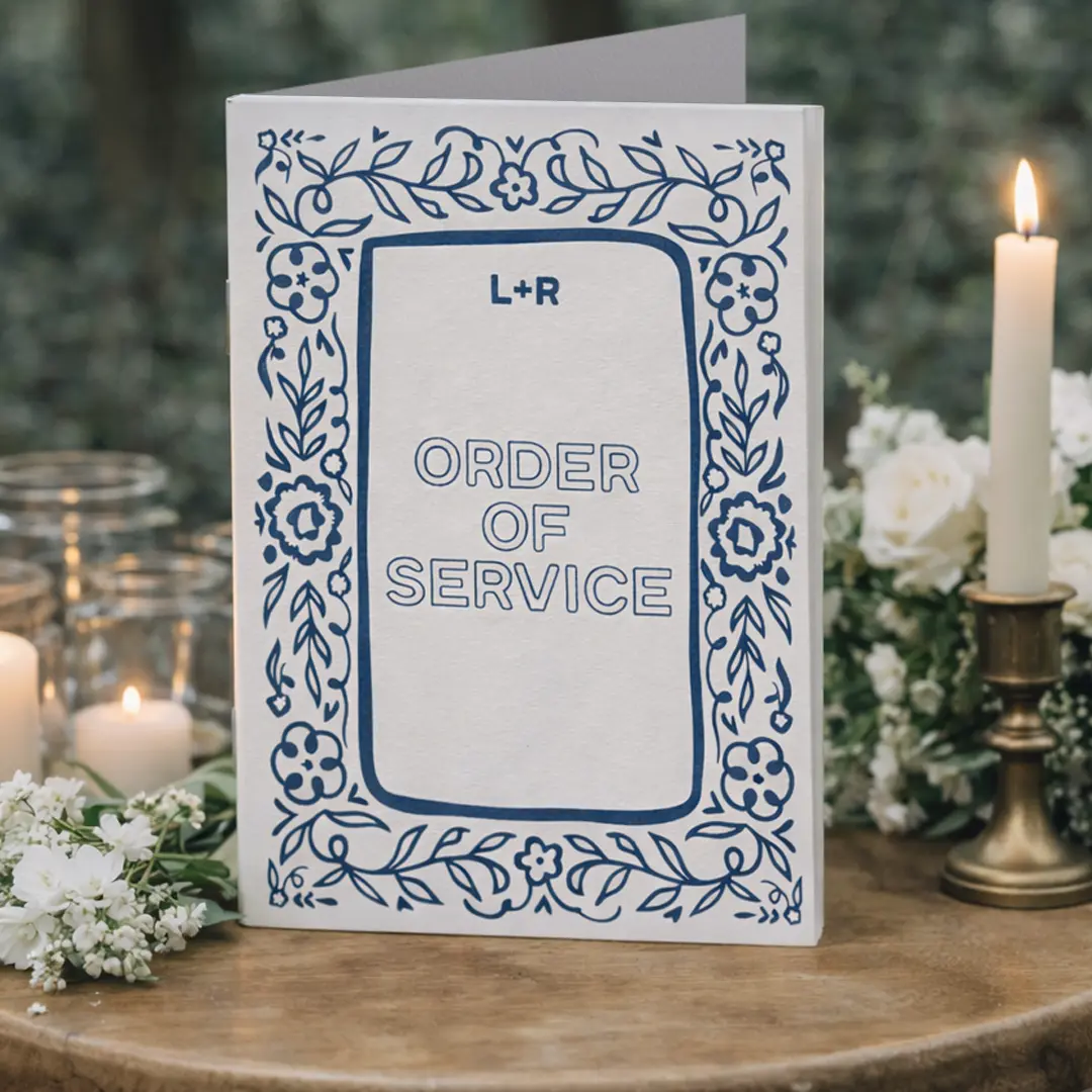 A white Folded Wedding Order of Service with blue floral detailing and the initials “L + R” on the cover, styled on a wooden surface surrounded by soft candlelight and white flowers