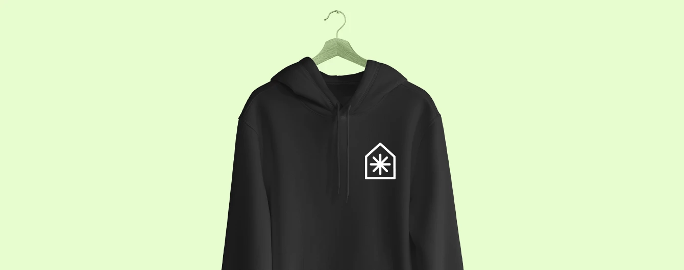 A black overhead hoodie hanging on a wooden coat hanger against a pale green background, featuring a white campaign logo of a simplified house containing an asterisk on the left chest.