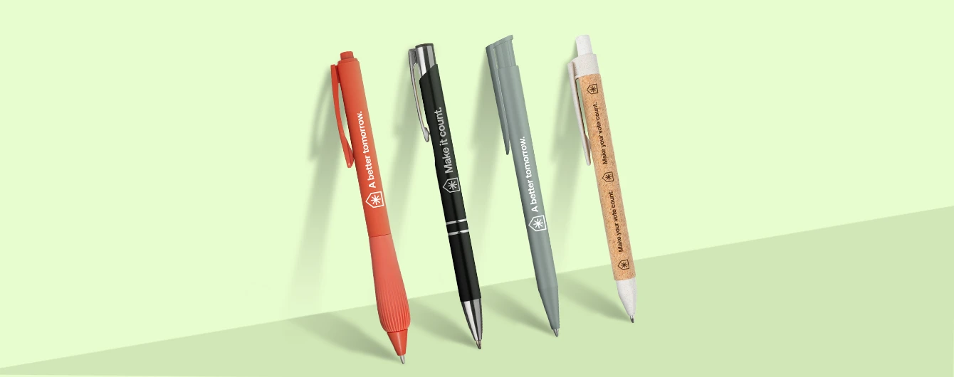 A row of four promotional pens in various styles and colours—orange, black, grey, and eco-friendly cork—leaning against a pale green wall, each printed with political campaign slogans such as "A better tomorrow" and "Make it count."