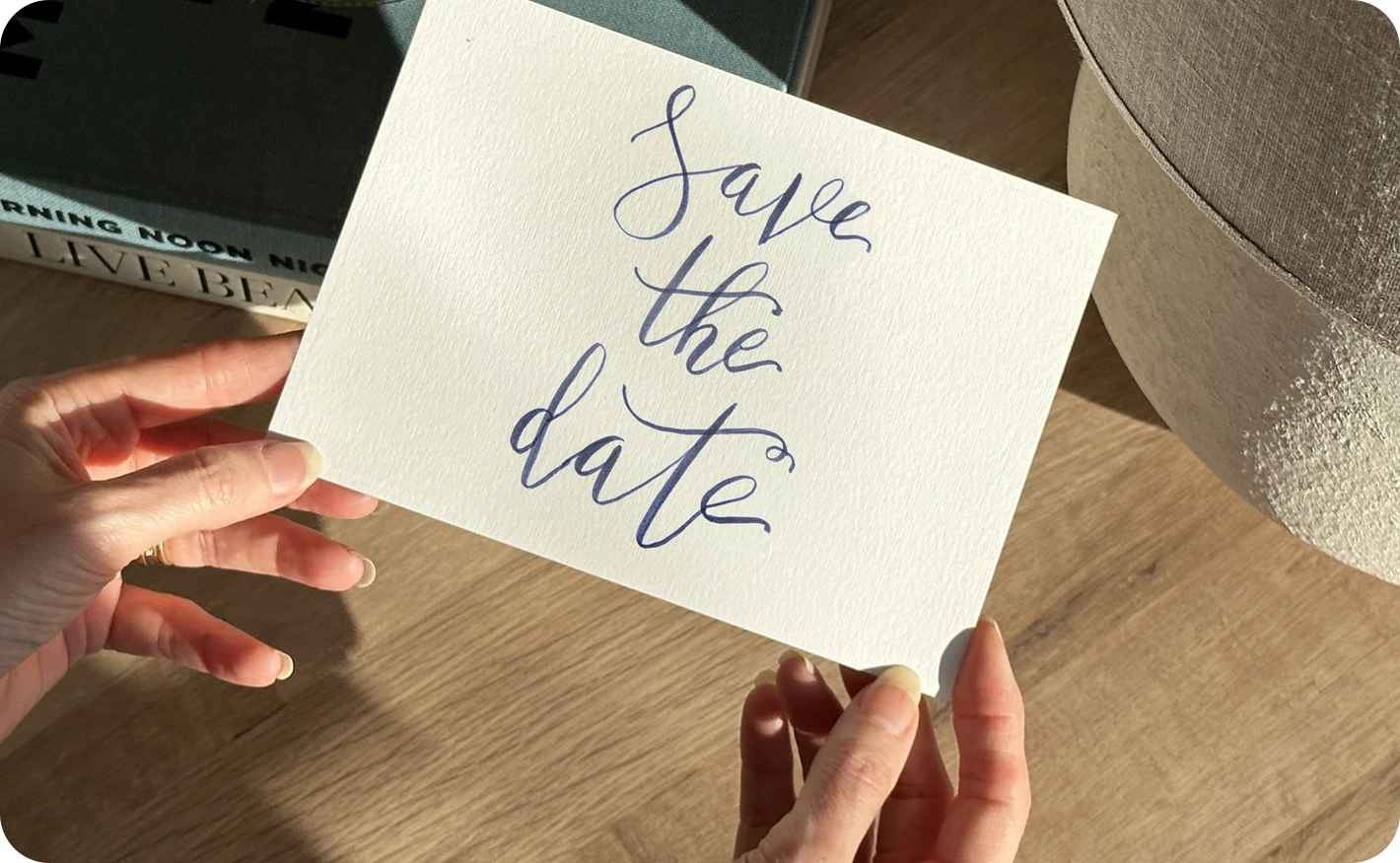 A hand holds a save the date card reading ‘Save the date’ above a wooden surface with a book and fabric in the background