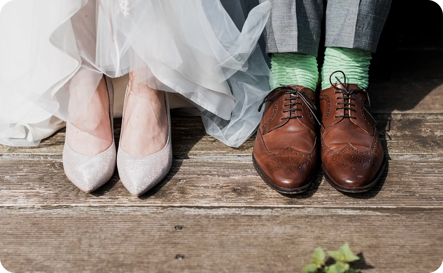 Close-up of a bride’s sparkly heels beside a groom’s brown dress shoes on a wooden surface