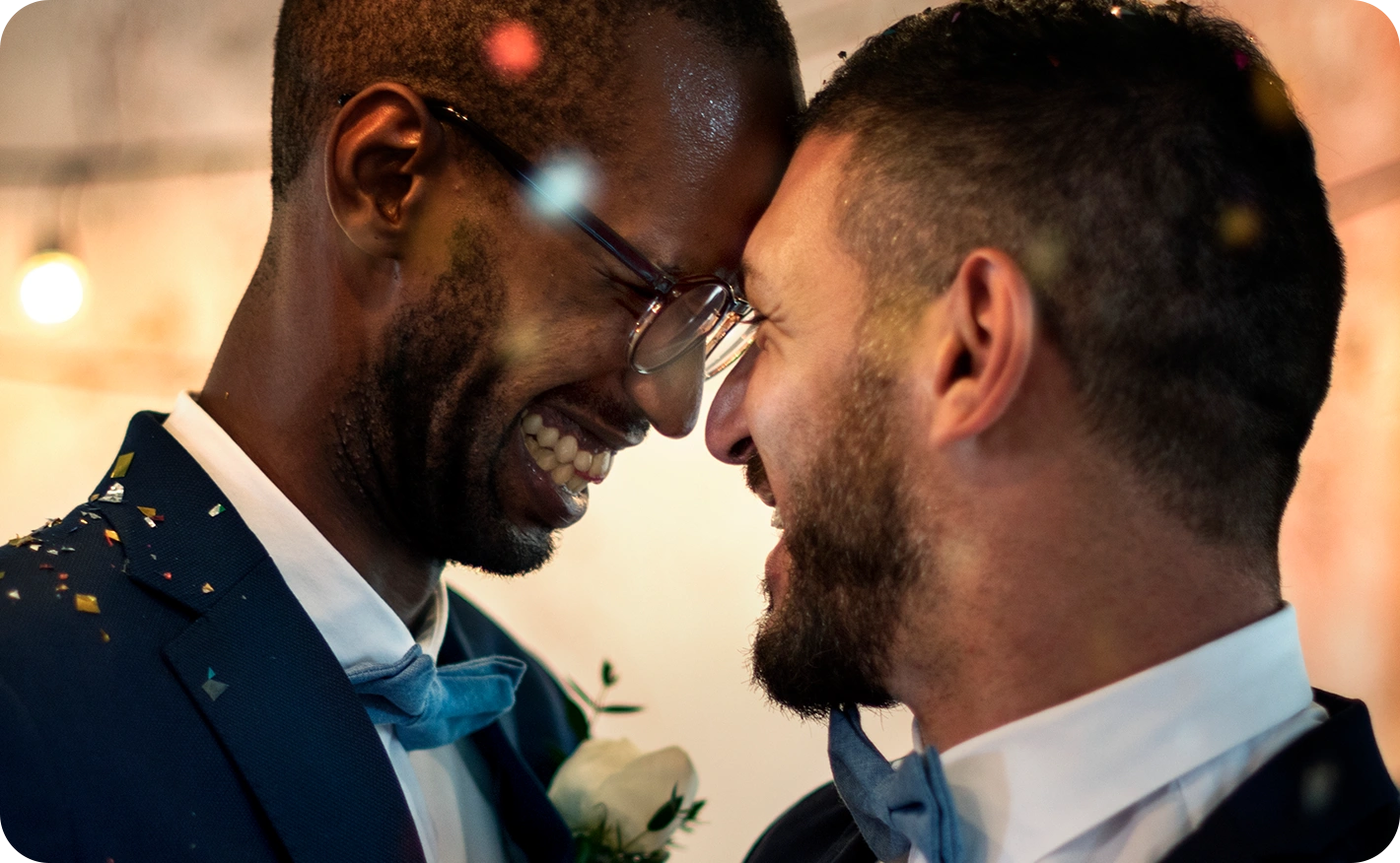 Two grooms in suits stand forehead to forehead, smiling as confetti falls around them in warm light at a wedding reception.