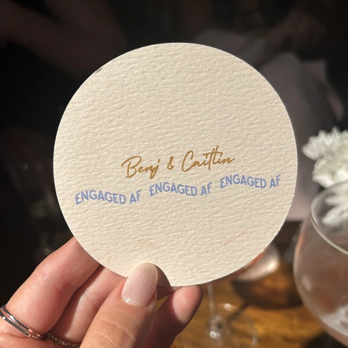 A hand holds a round textured paper Wedding Coaster printed with “Benj & Caitlin” in gold script above three stacked lines reading “ENGAGED AF” in bold blue lettering