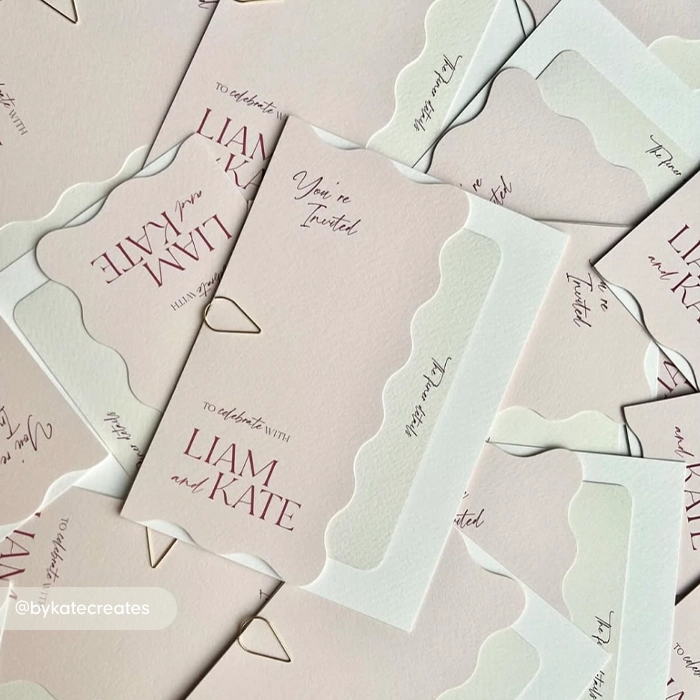 A scattered arrangement of pastel Wedding Invitations with wavy cut‑out edges. Each card reads ‘You’re invited to celebrate with Liam and Kate.’