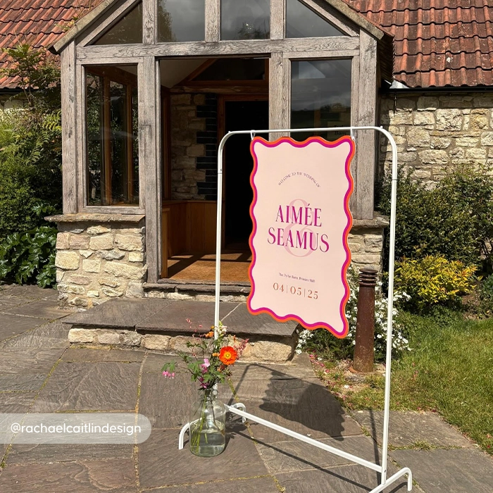 An outdoor wavy Wedding Welcome Sign on a white metal stand in front of a rustic stone building. The pink sign with a wavy red border reads ‘Welcome to the wedding of Aimée & Seamus, 04 | 05 | 23.’