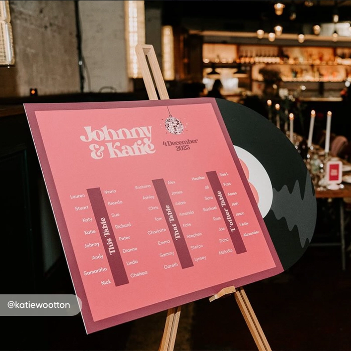 A retro‑style Wedding Table Plan Board on an easel with a pink vinyl‑record design, titled ‘Johnny & Katie, 4 December 2023,’ showing three guest‑list columns labelled ‘This Table,’ ‘That Table,’ and ‘Other Table’.