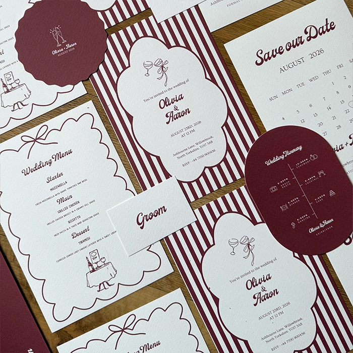 “Coordinated burgundy‑and‑white wedding stationery set displayed on a pink background, including an invitation, save‑the‑date card, menu, itinerary, and a place card labelled ‘Groom,’ all personalised for Olivia and Aaron