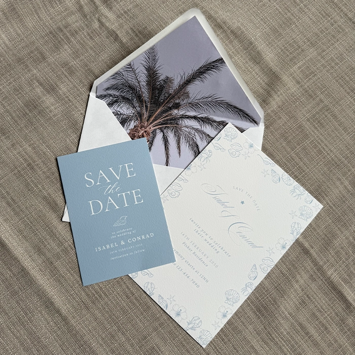 Blue Save the Date Card and white illustrated Wedding Invitation displayed on a textured fabric surface, with an Envelope Liner with a palm‑tree photo behind them.