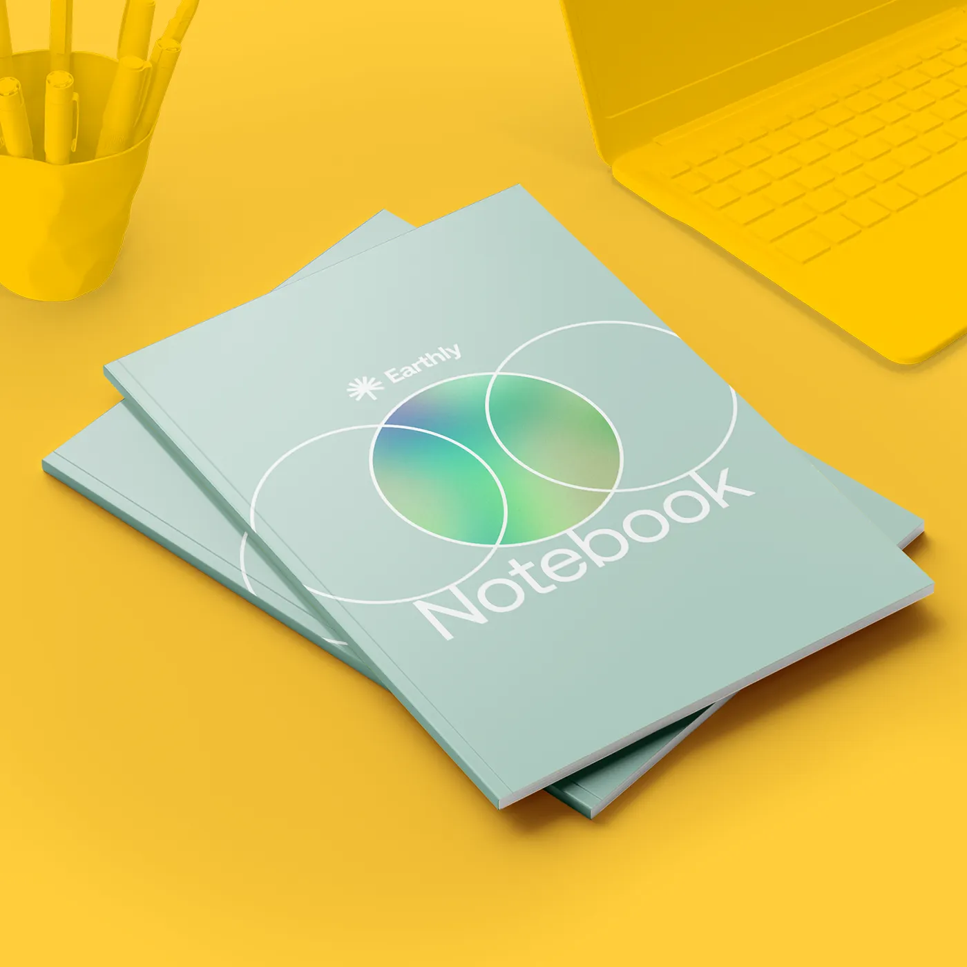 Two pastel‑coloured Perfect Bound Notebooks stacked next to a laptop and a pot of stationery.