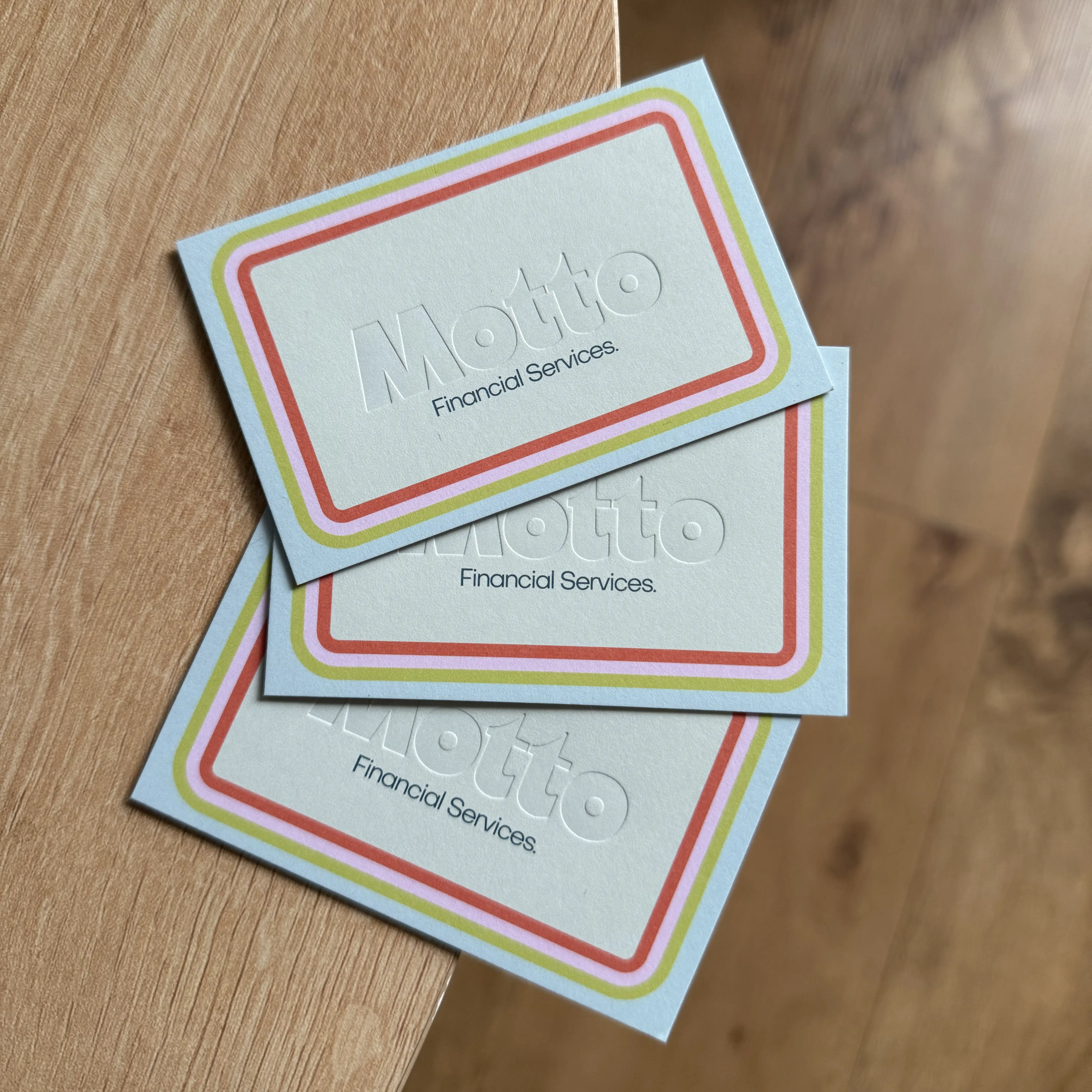 A fanned-out trio of pale mint business cards for "Motto Financial Services" rests on a wooden surface, featuring the "Motto" logo deeply debossed into the centre, framed by a vibrant, multi-coloured border of orange, pink, and yellow lines.