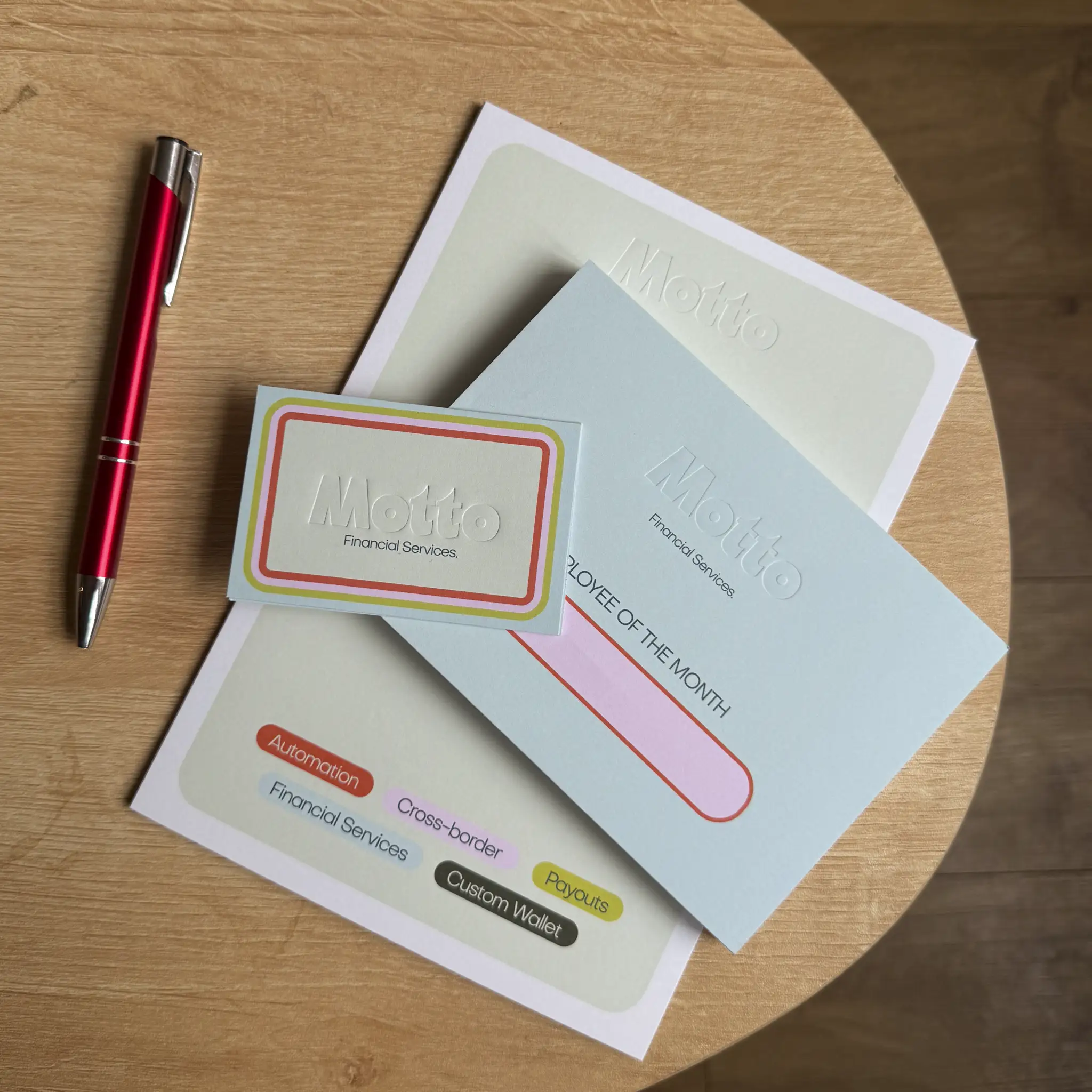 A red pen lies next to a collection of "Motto Financial Services" stationery on a wooden table, featuring a certificate and business cards with the company name "Motto" deeply pressed into the thick, pale mint and light blue cardstock using a tactile debossing technique.
