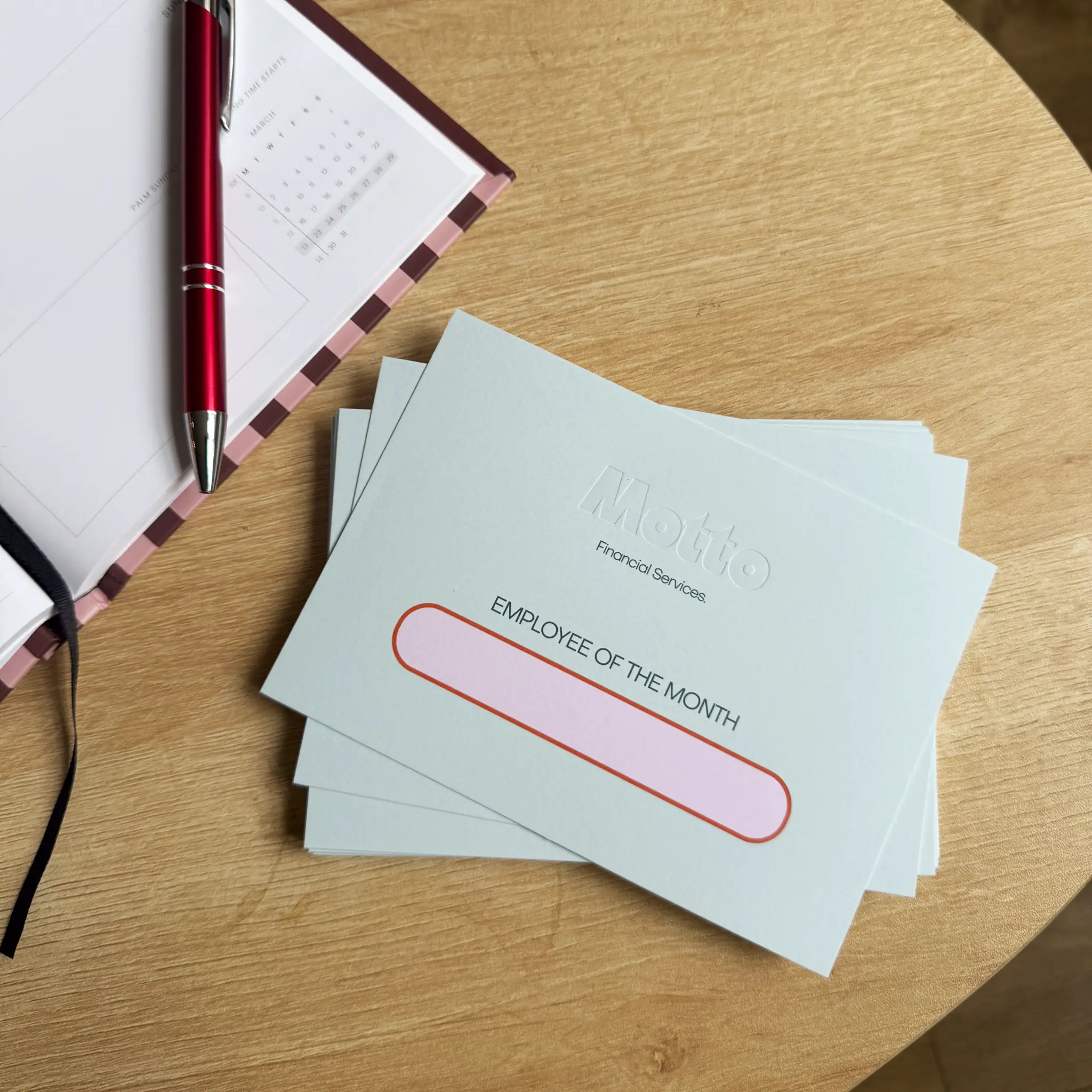 A stack of pale mint "Employee of the Month" cards rests on a wooden desk next to an open planner, featuring the "Motto Financial Services" logo deeply pressed into the centre of the thick cardstock using a crisp, inkless debossing technique.