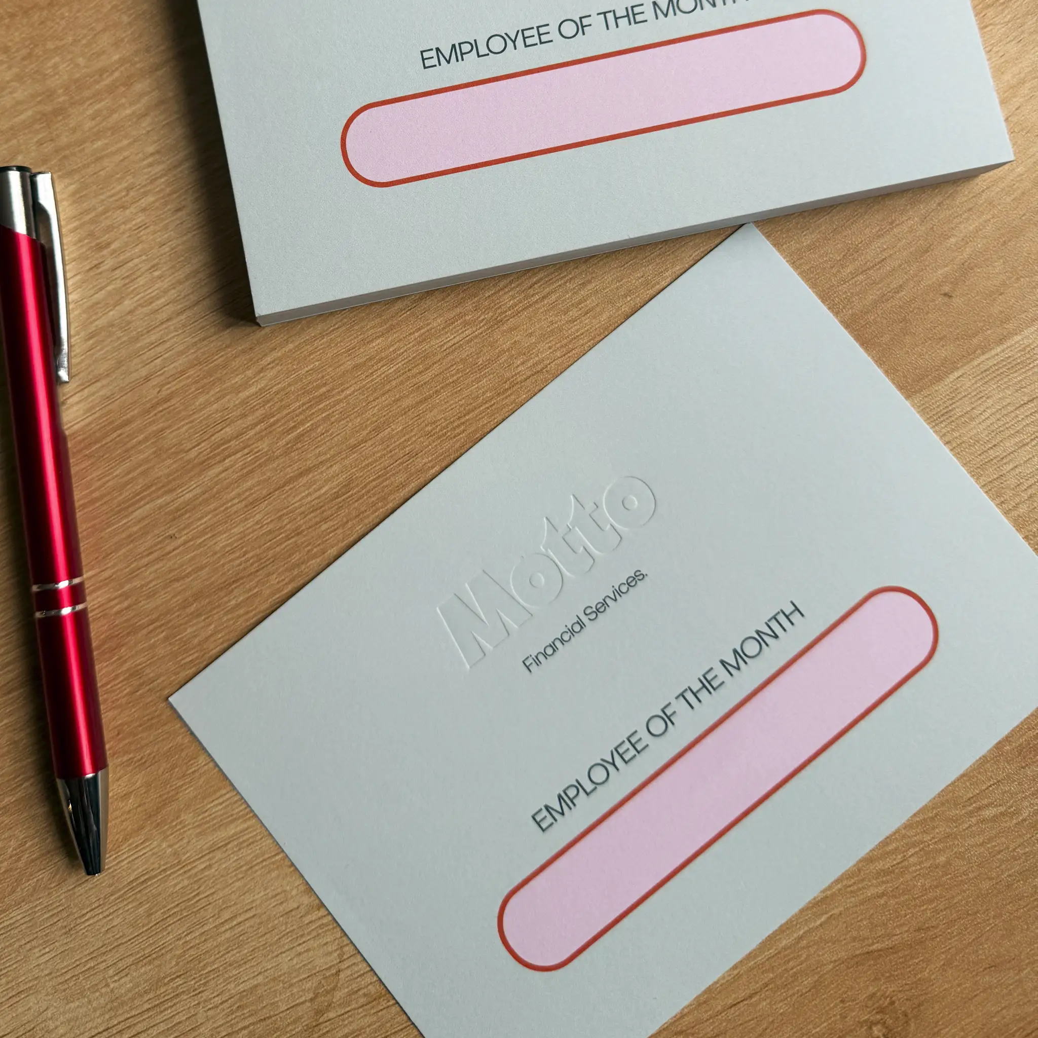 A close-up shows a red pen next to a pale mint "Employee of the Month" card, featuring the "Motto Financial Services" logo deeply debossed into the centre, creating a tactile, inkless impression that catches the light and casts soft shadows on the thick cardstock.