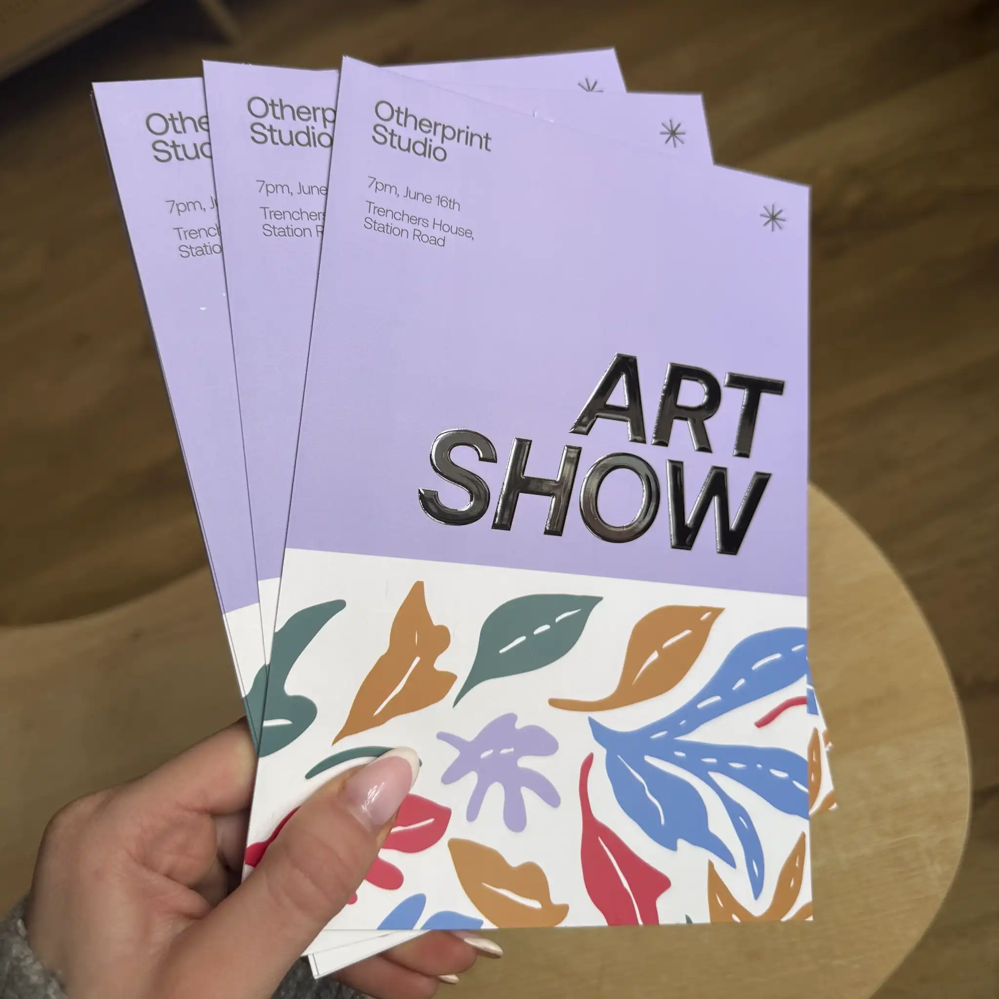 A person holds a fan of three lavender and white "Art Show" flyers, where the main title is highlighted with a thick, glossy spot UV finish that gives the black lettering a high-shine, liquid appearance against the matte paper.