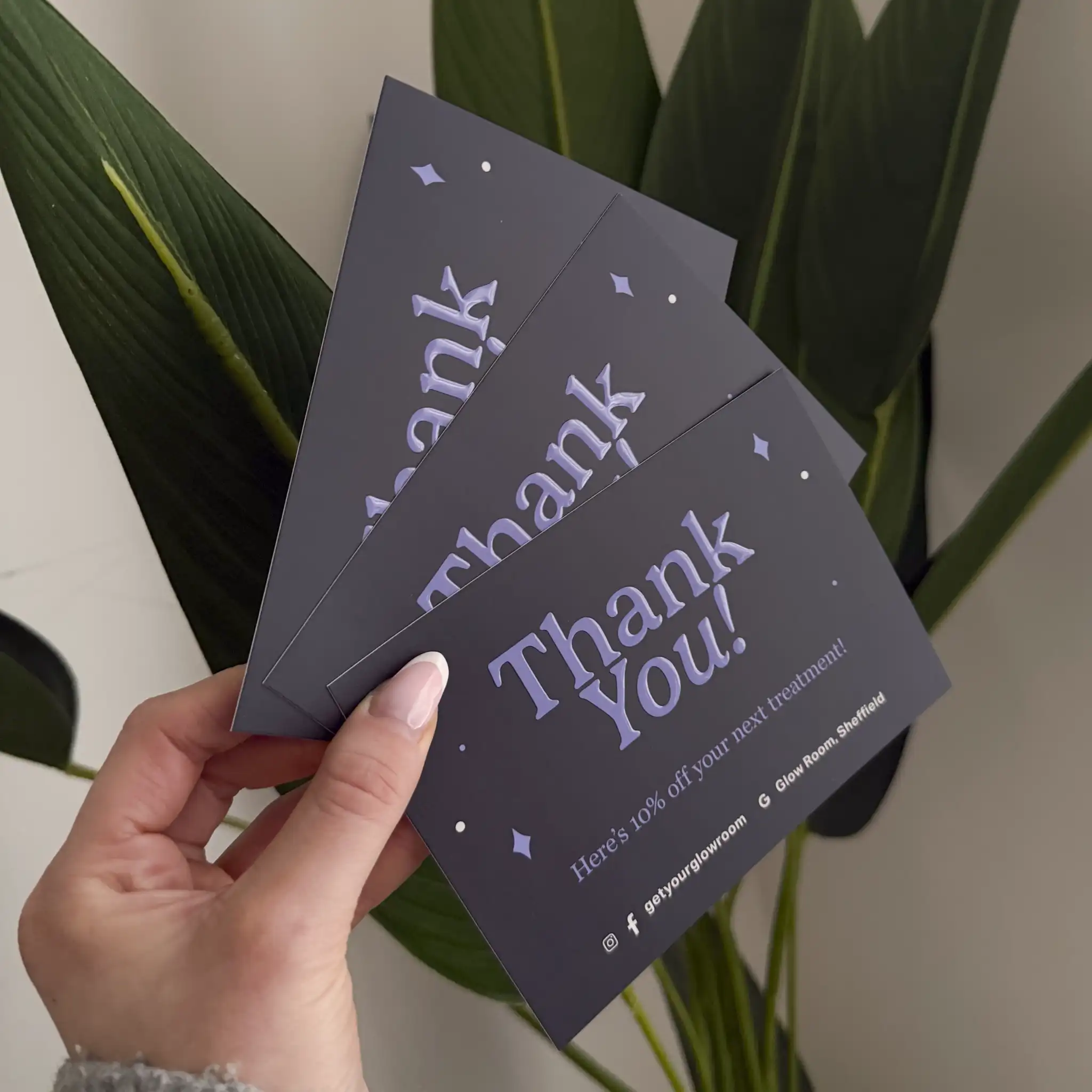 A hand holds a fan of three dark purple "Thank You!" postcards, showcasing a glossy, raised spot UV finish on the large typography and small star accents that creates a tactile, high-shine contrast against the matte cardstock.