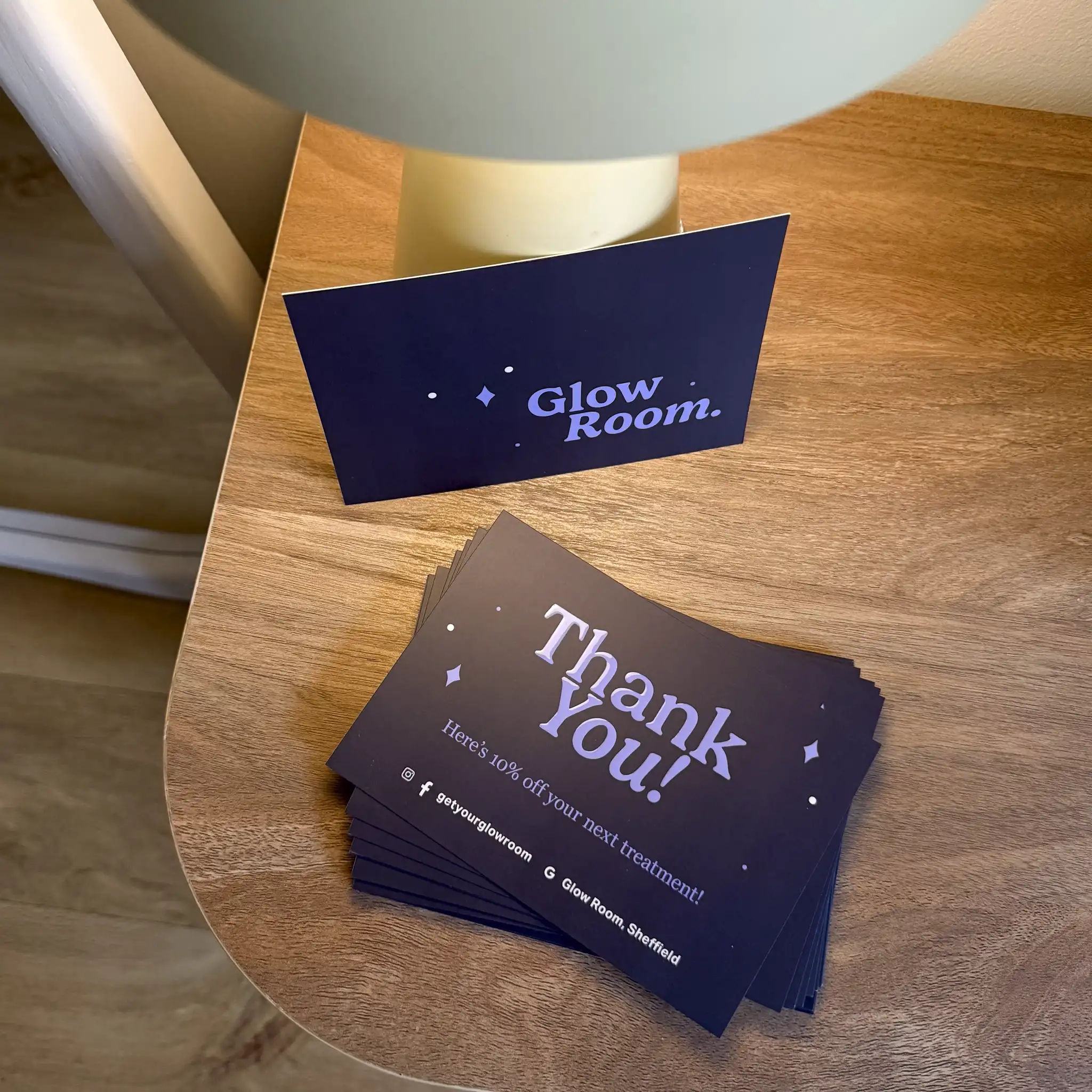 A stack of dark navy postcards for "Glow Room" sits on a wooden surface, featuring the words "Thank You!" and small star motifs finished with a glossy, raised spot UV coating that catches the light.