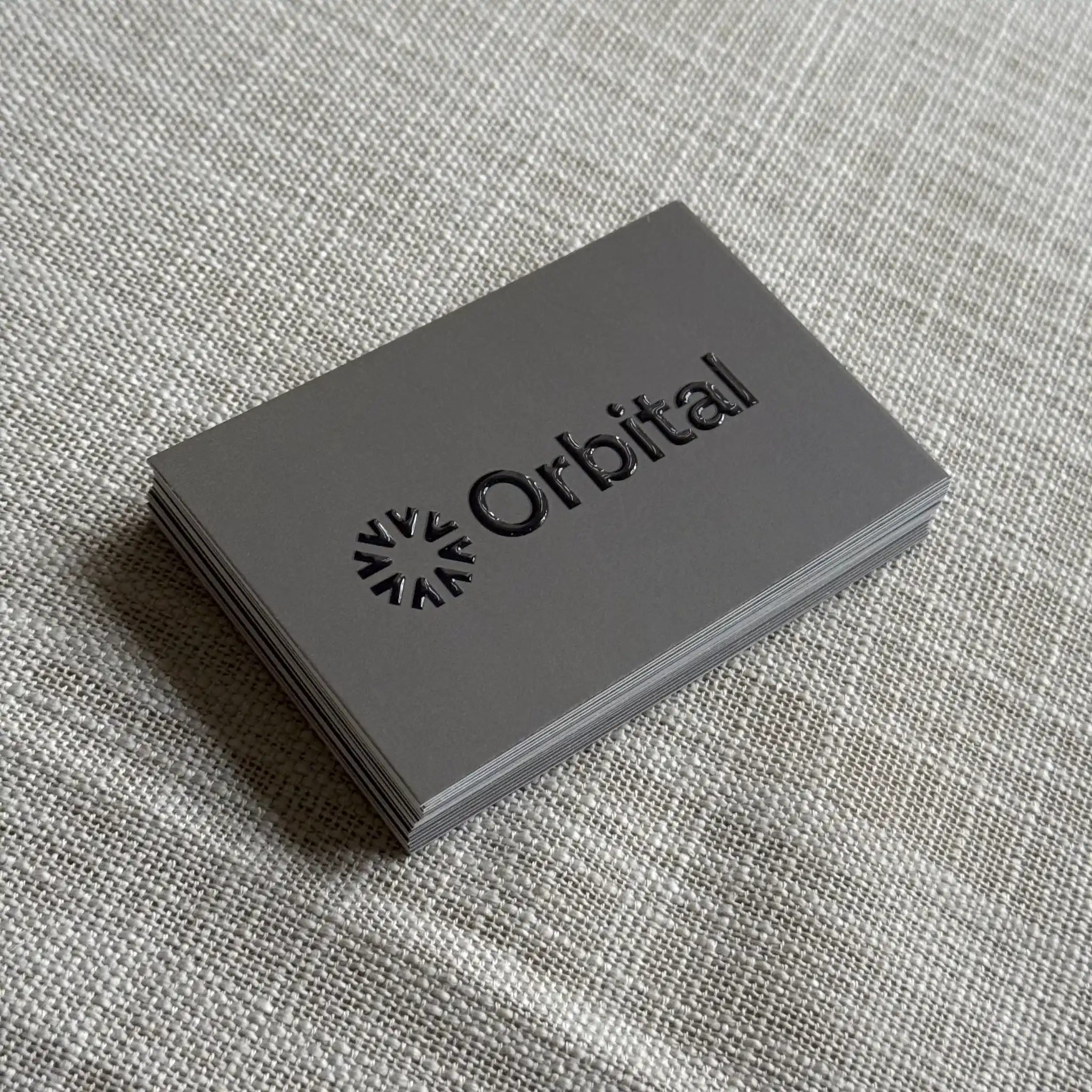 A stack of matte grey business cards for "Orbital" features a minimalist sun-like logo and company name finished in a raised, high-gloss spot UV that creates a striking 3D texture against the flat background.