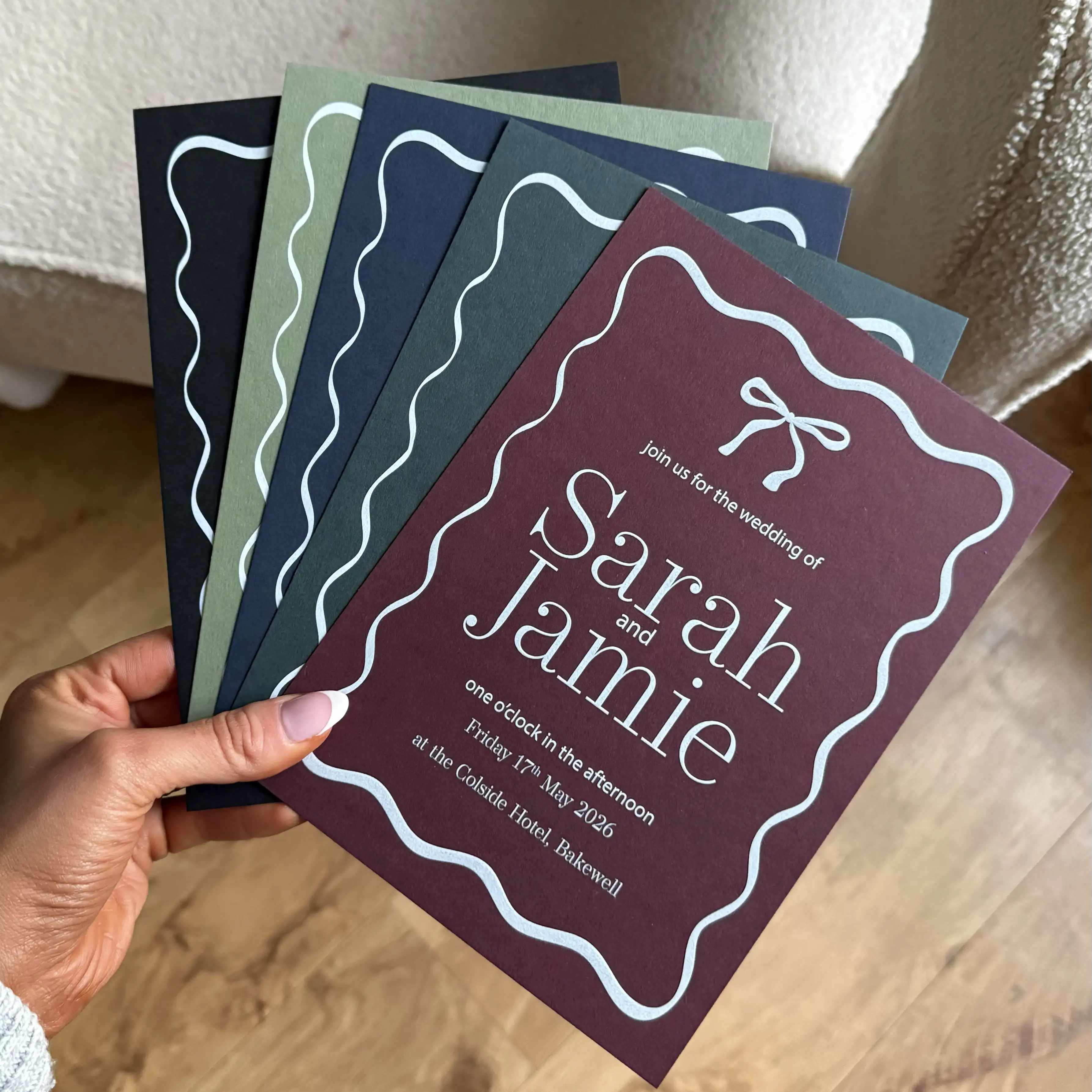 A hand holds a fanned-out selection of wedding invitations in deep burgundy, forest green, navy, and black, each featuring a wavy white border, a delicate bow illustration, and clean white-ink typography for a modern, high-contrast finish.