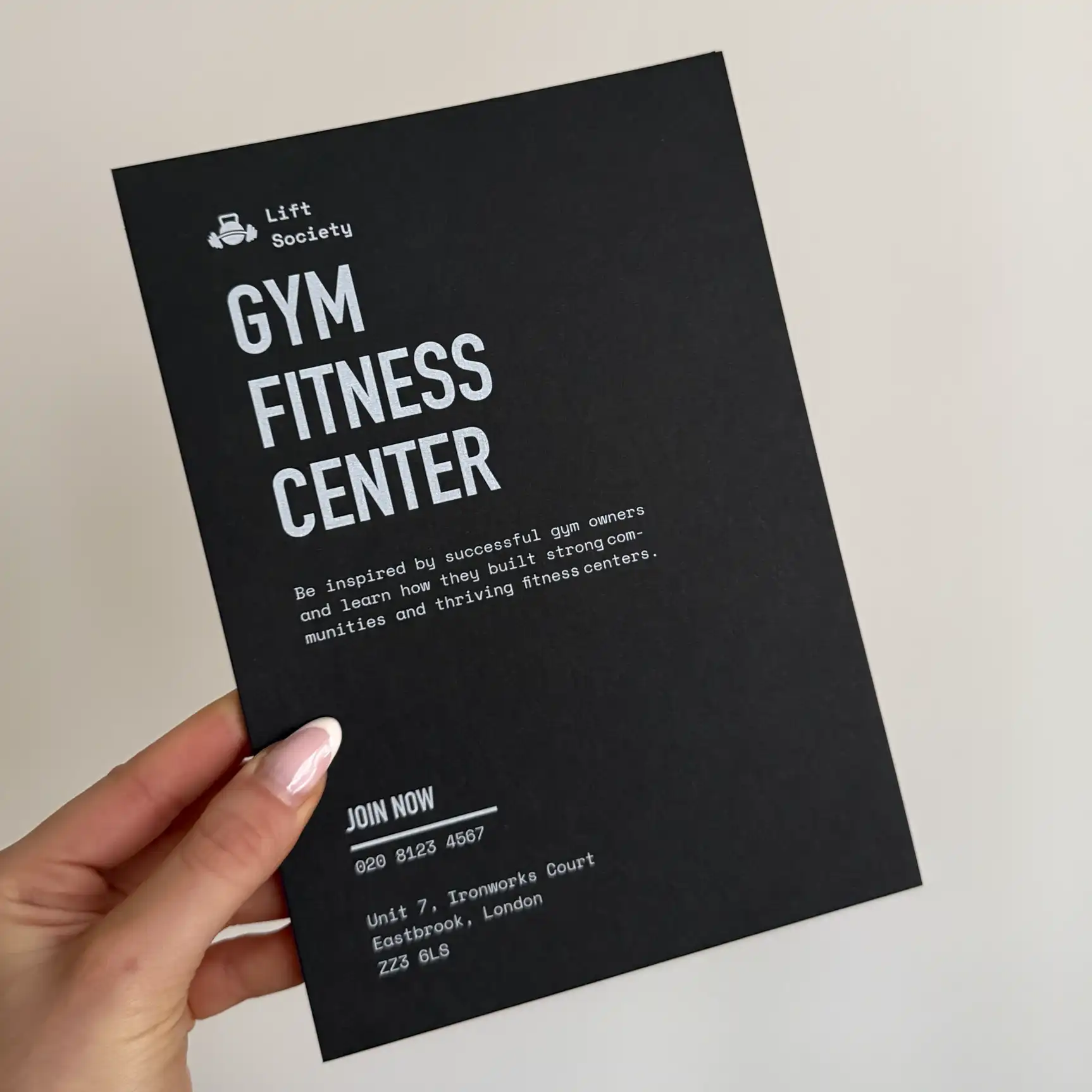 A person holds a black "Gym Fitness Center" flyer for Lift Society, featuring bold, minimalist white ink typography and a small kettlebell icon that stands out sharply against the dark cardstock.