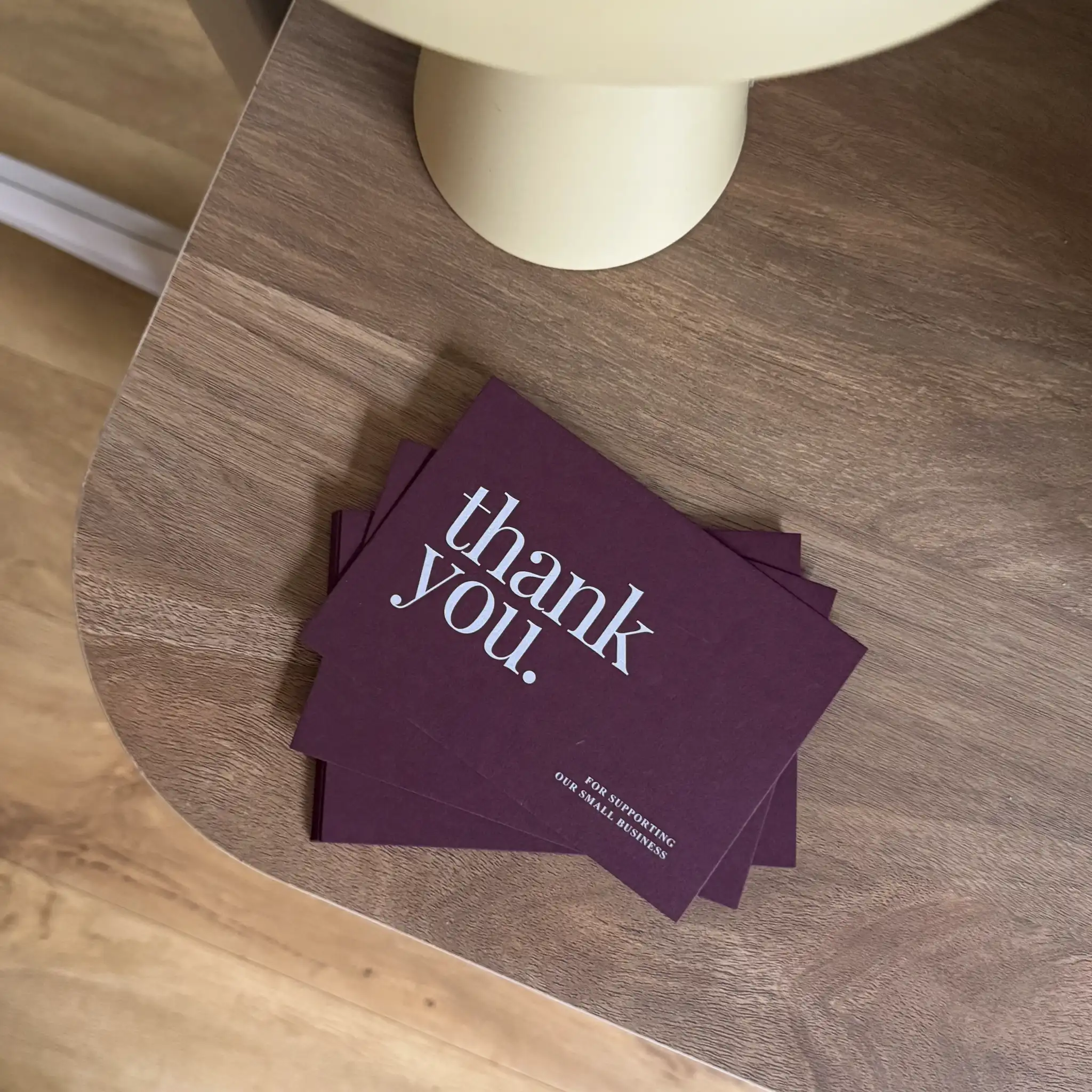 A small stack of deep plum-coloured "thank you" cards rests on a wooden table, featuring minimalist white ink lettering in a clean serif font that provides a sharp, crisp contrast against the rich purple cardstock.