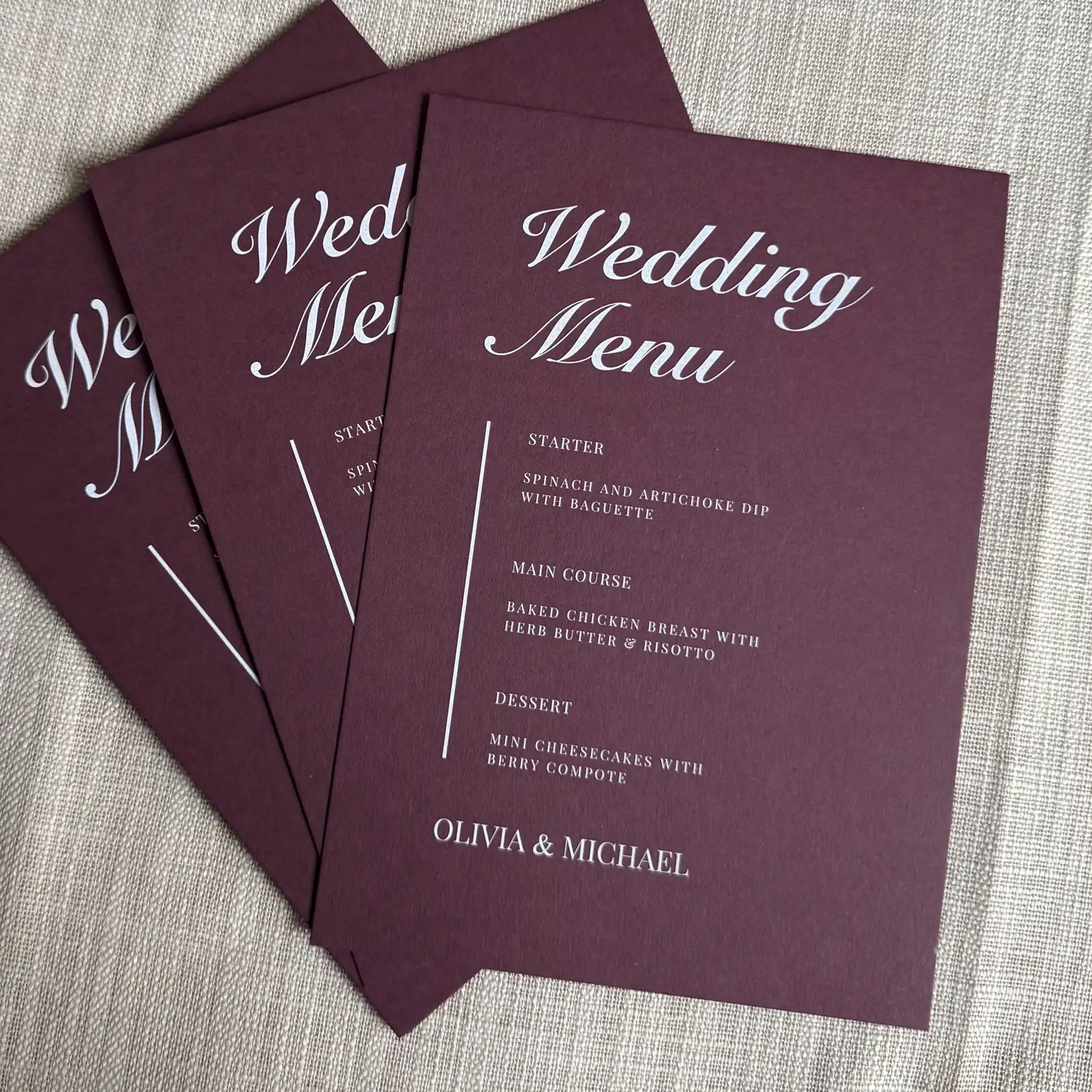 A fanned-out trio of burgundy wedding menus features elegant white ink printing, with "Wedding Menu" in a large, sweeping script at the top and the names "Olivia & Michael" at the bottom, flanking a clean, minimalist list of courses.
