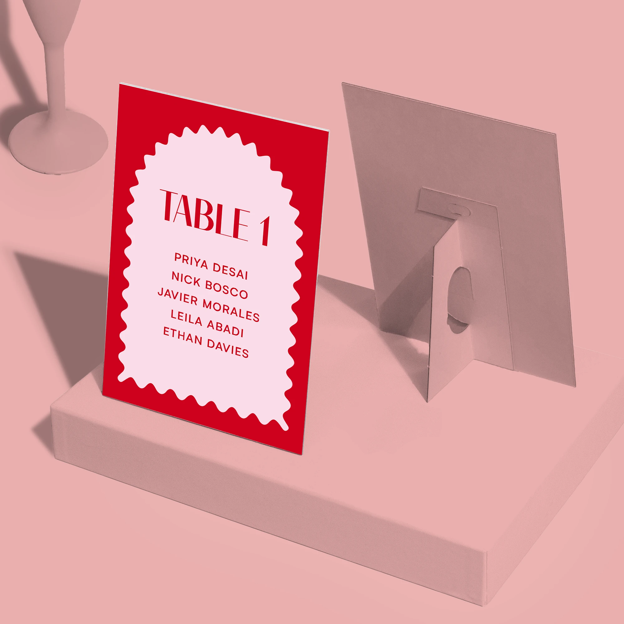 A red and pink Strut Card labeled "TABLE 1" with a list of guest names, shown from the front and back to display the easel stand.
