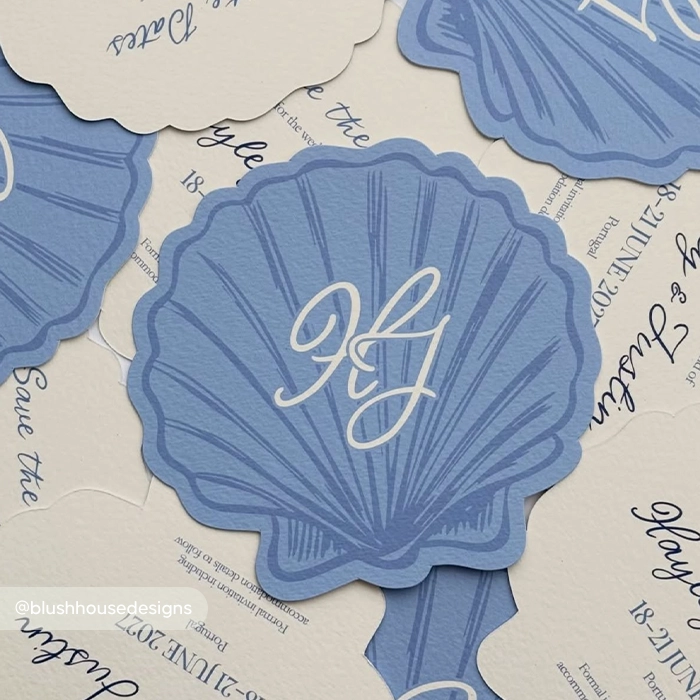 Blue scalloped seashell-shaped wedding stationery featuring an elegant cream-coloured cursive monogram.