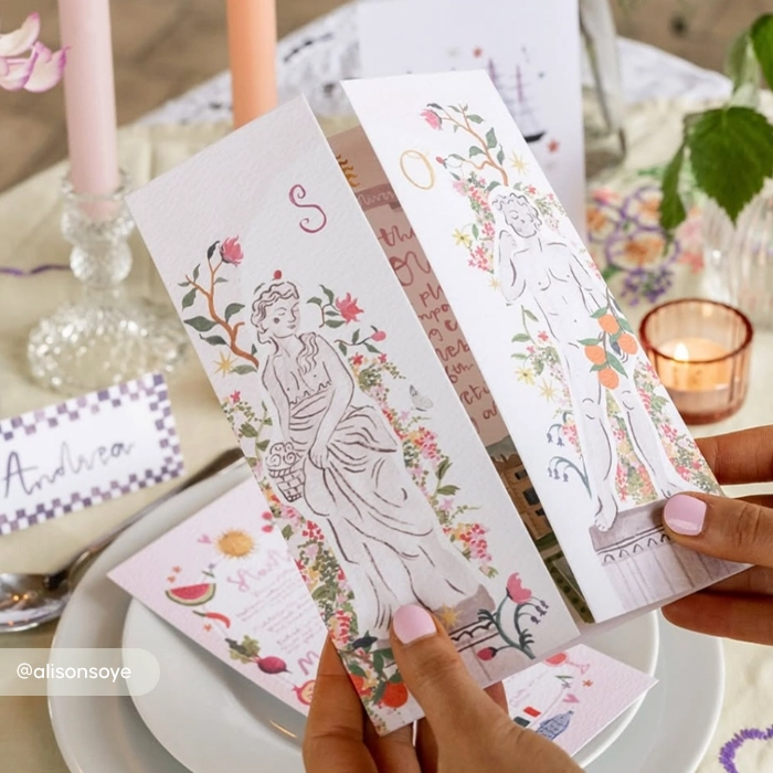 A pair of hands holds open a Gatefold Wedding Invitation featuring whimsical illustrations of Greek-style statues adorned with colourful flowers and fruit.
