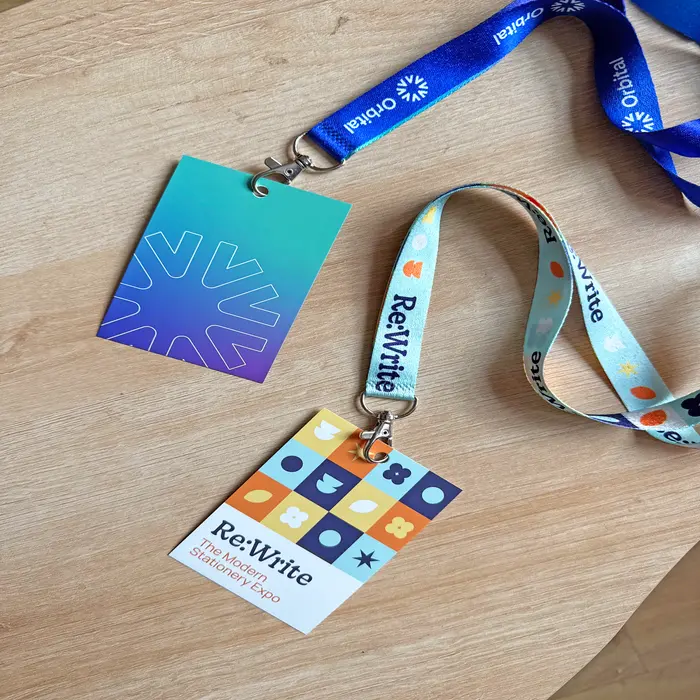Two event Lanyards rest on a light wooden surface: one purple-to-teal gradient Lanyard Card with a logo and a branded strap, and another card for "Re:Write - The Modern Stationery Expo" with a colourful geometric pattern and matching light blue strap.