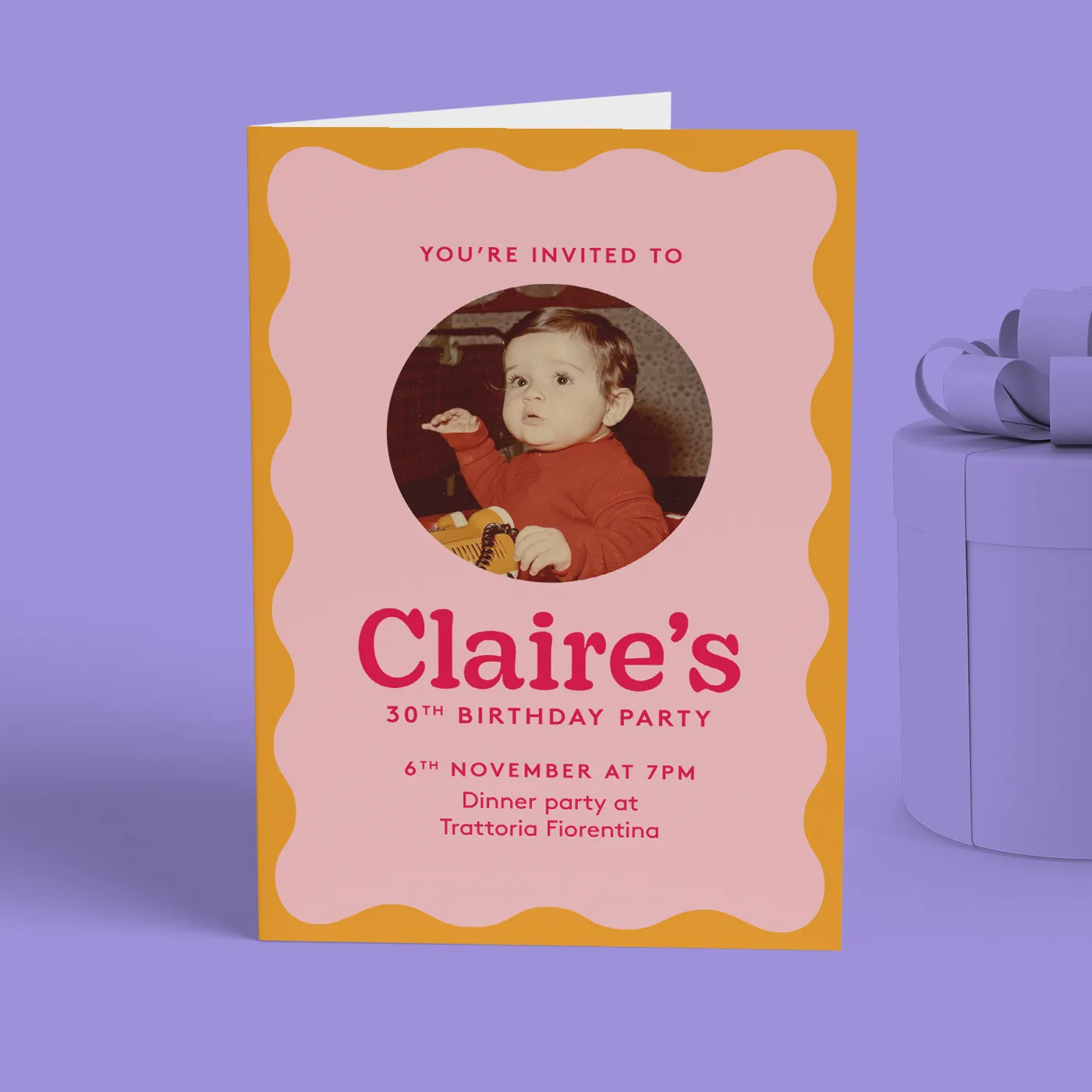 A folded birthday invitation standing beside a lavender gift box on a purple surface