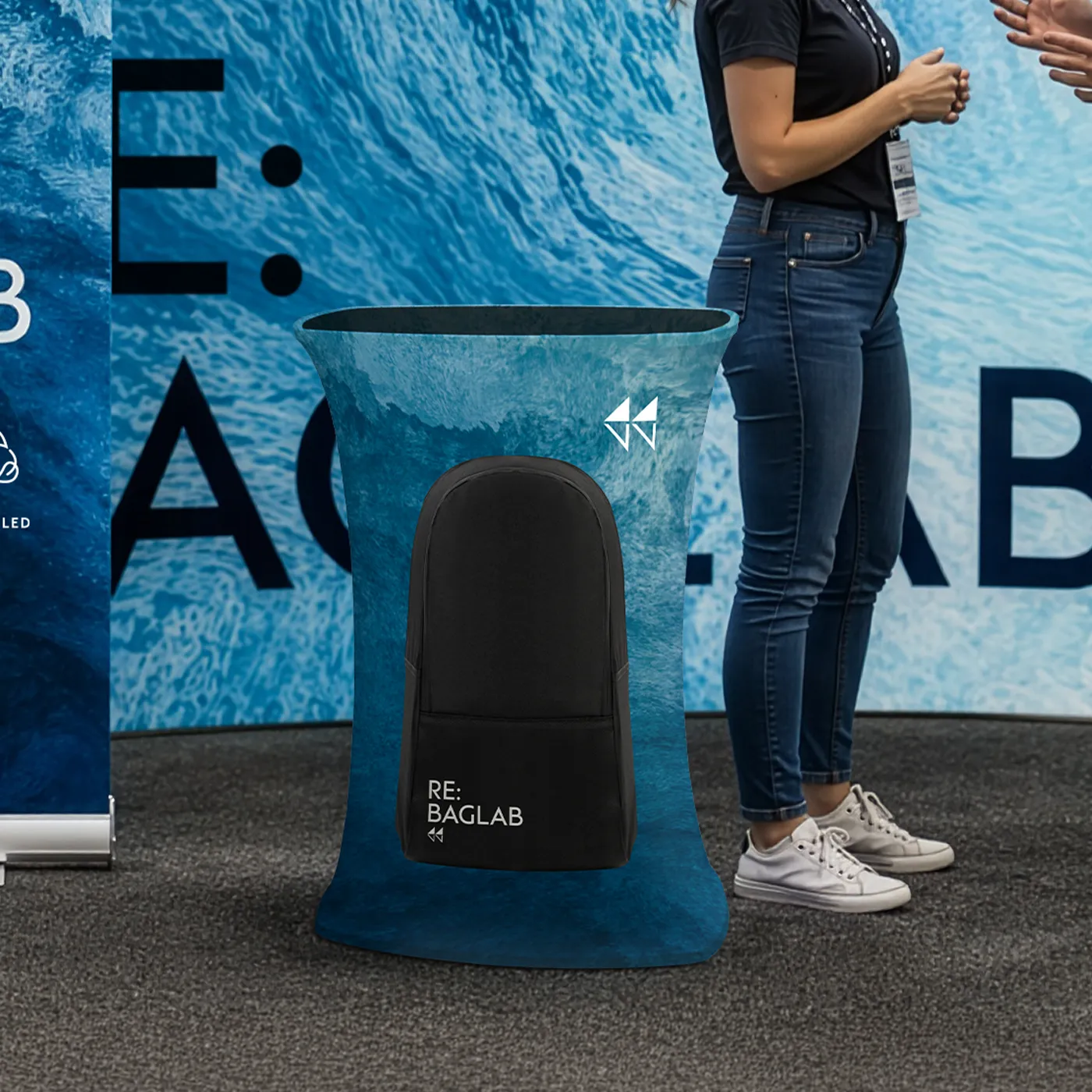 A Square Pop Up Counter at an exhibition stand for "RE: BAGLAB" featuring a blue ocean-textured graphic and a black backpack displayed in the centre.