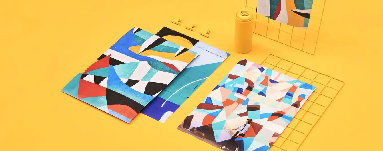 Two vibrant art prints featuring bold, colourful geometric patterns and abstract shapes, arranged flat on a bright yellow surface alongside a yellow spray can and metal wire grids.